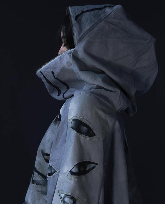 Profile of a person wearing a sculptural grey hooded garment with eye-shaped cut-outs