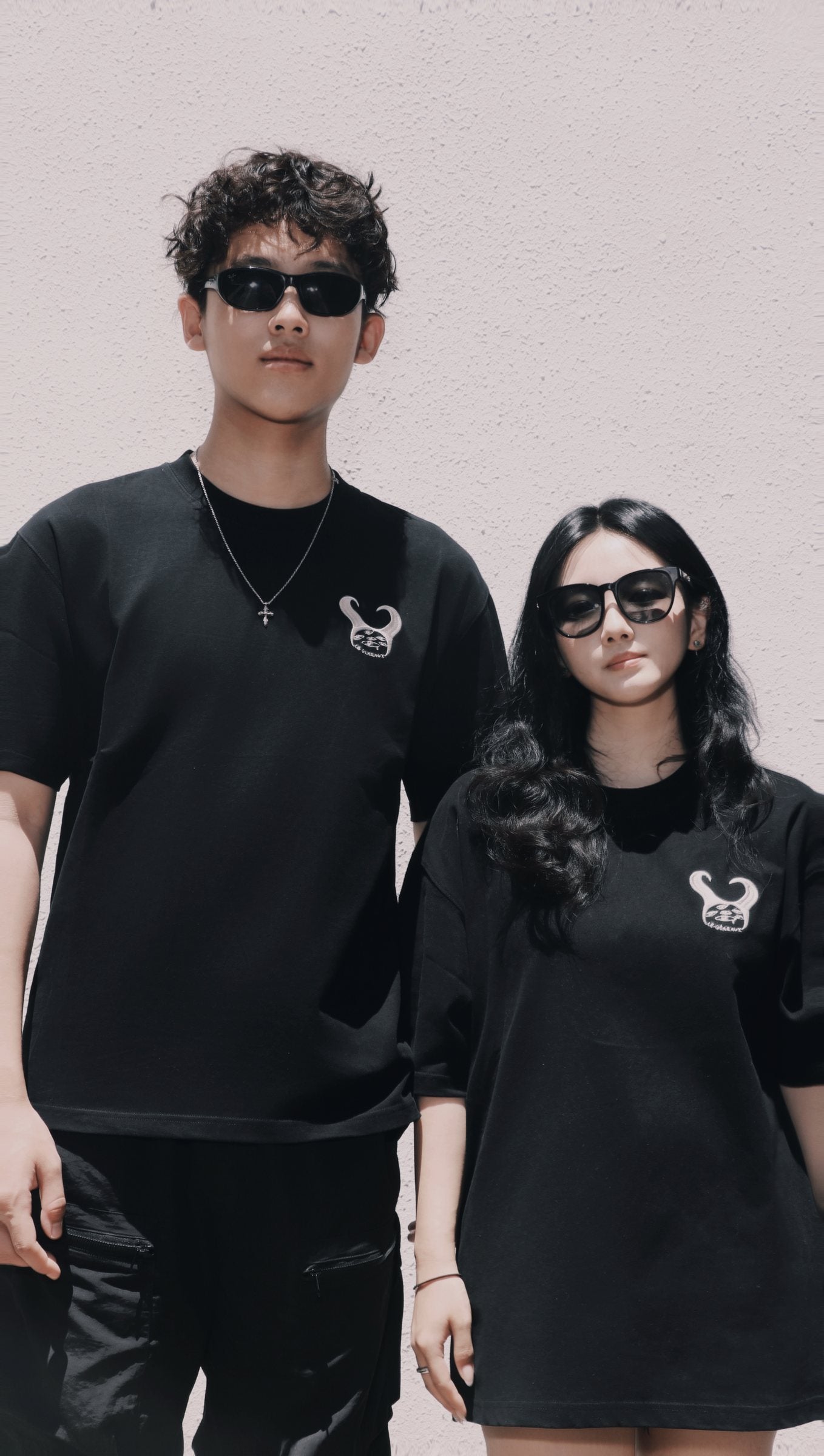 Two people wearing black LE GÉMEAUX T-shirts and sunglasses against a pale textured wall.
