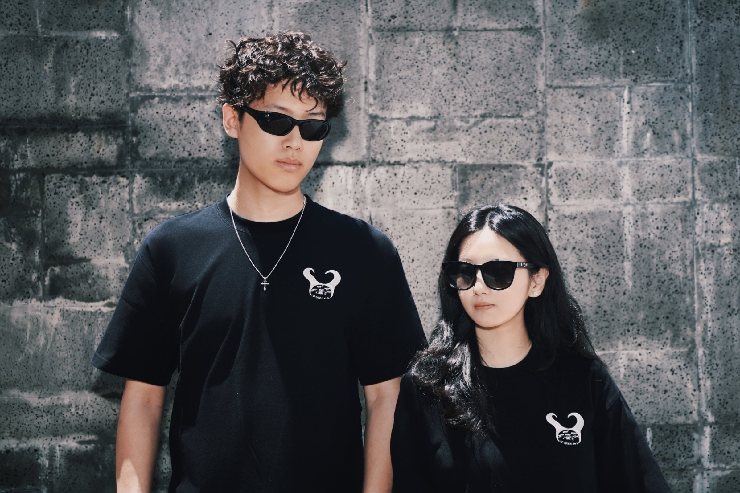 Two people in black graphic T-shirts and dark sunglasses standing against a textured grey block wall.