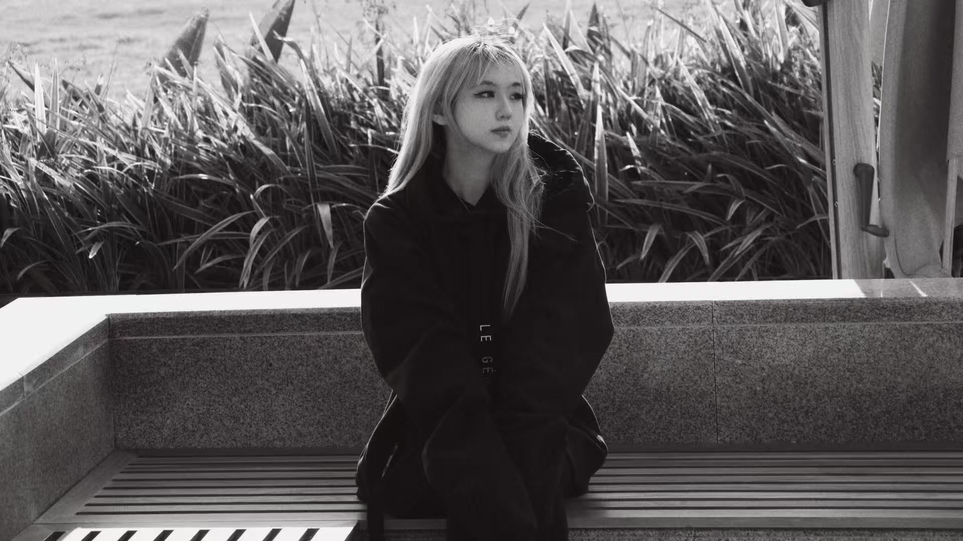Person wearing a black LE GÉMEAUX hoodie seated on a bench in front of tall plants.