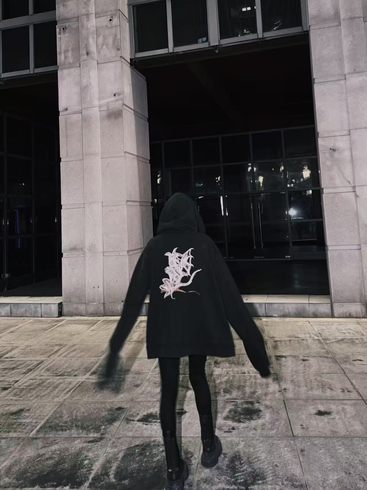 Rear view of a person in a black hoodie with a large pale back graphic, walking toward a concrete building.