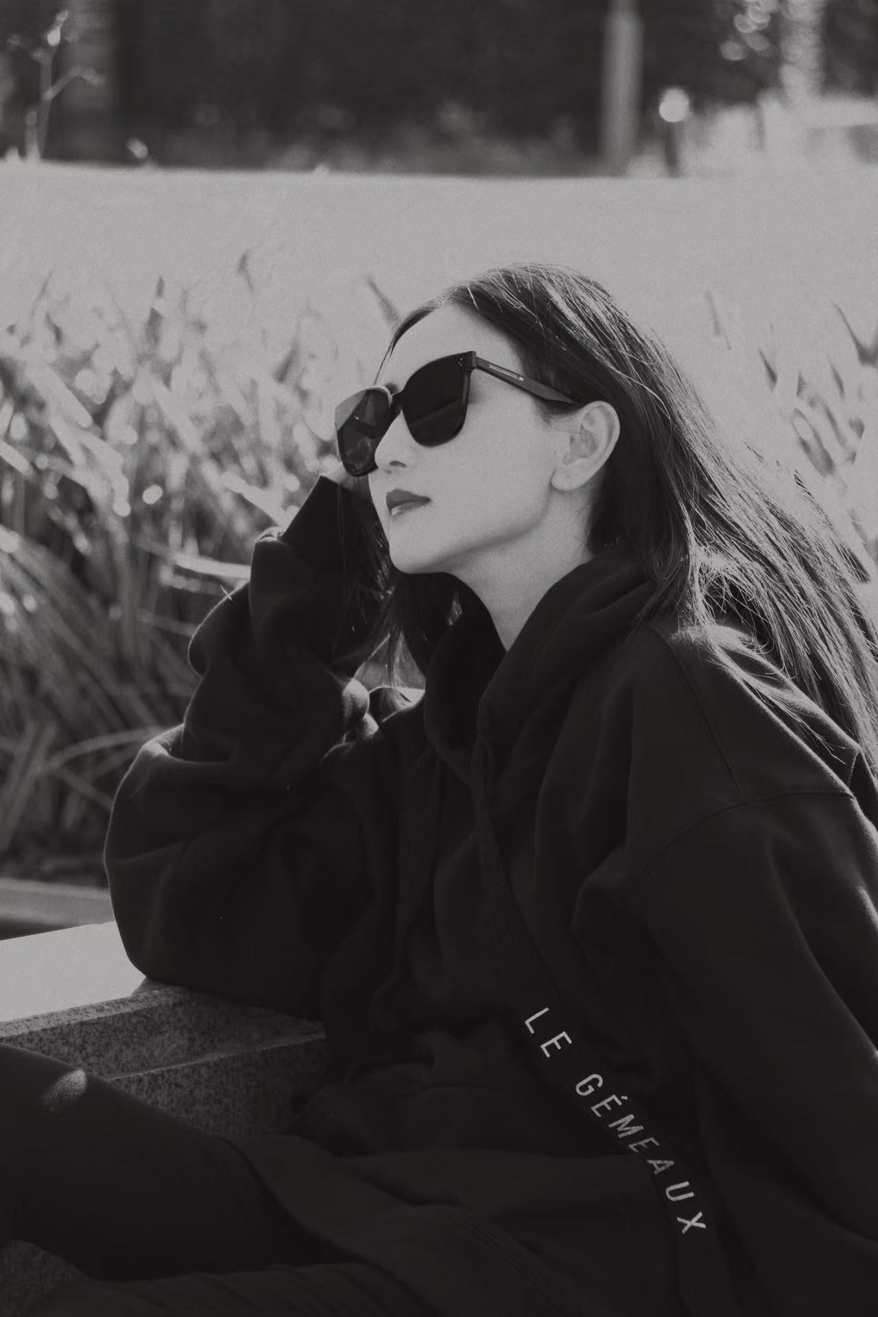 Black-and-white portrait of a person in dark sunglasses and an oversized black hoodie with LE GÉMEAUX lettering on the sleeve.