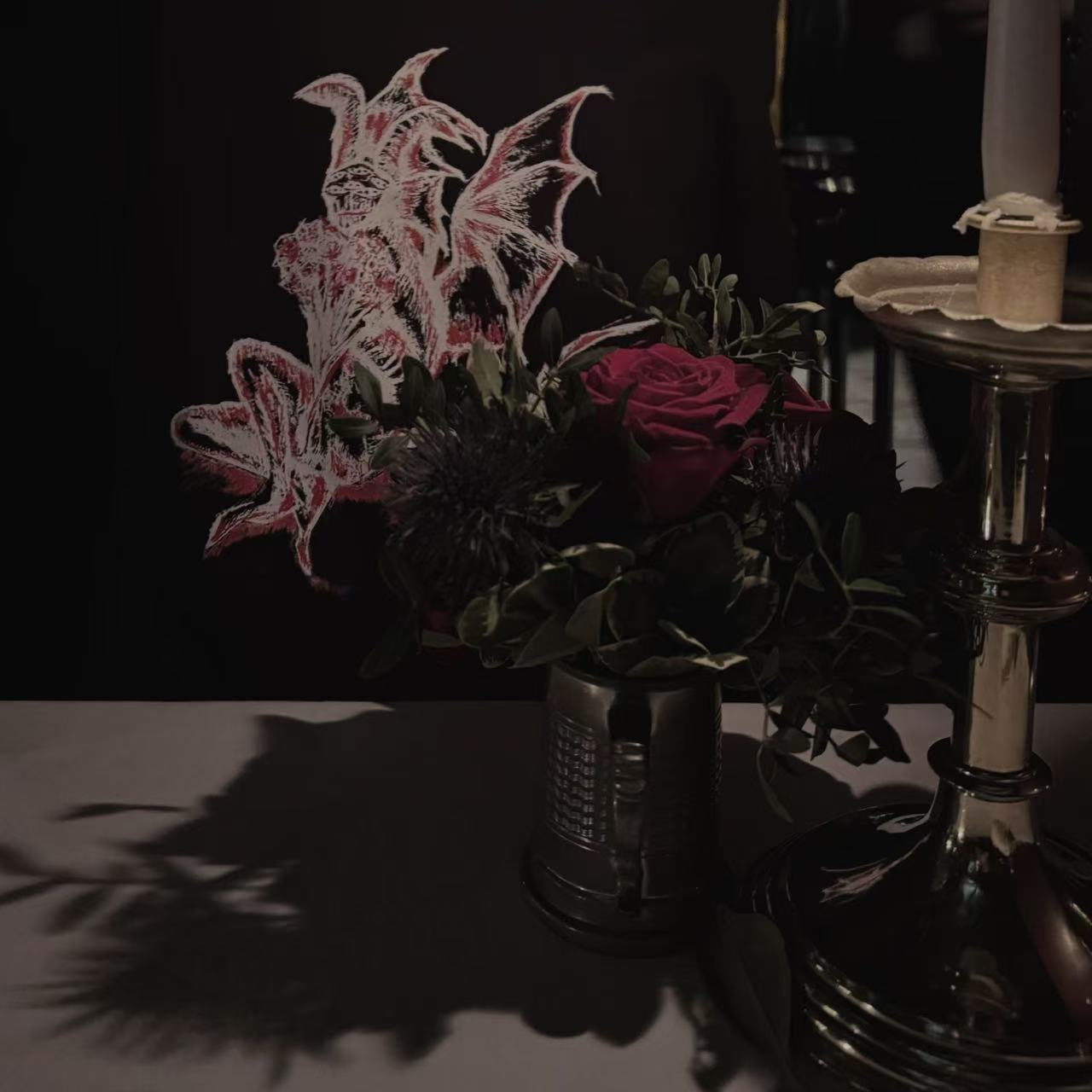 A dark floral arrangement with red roses beside a tall candleholder, against a black backdrop with a pale winged illustration.