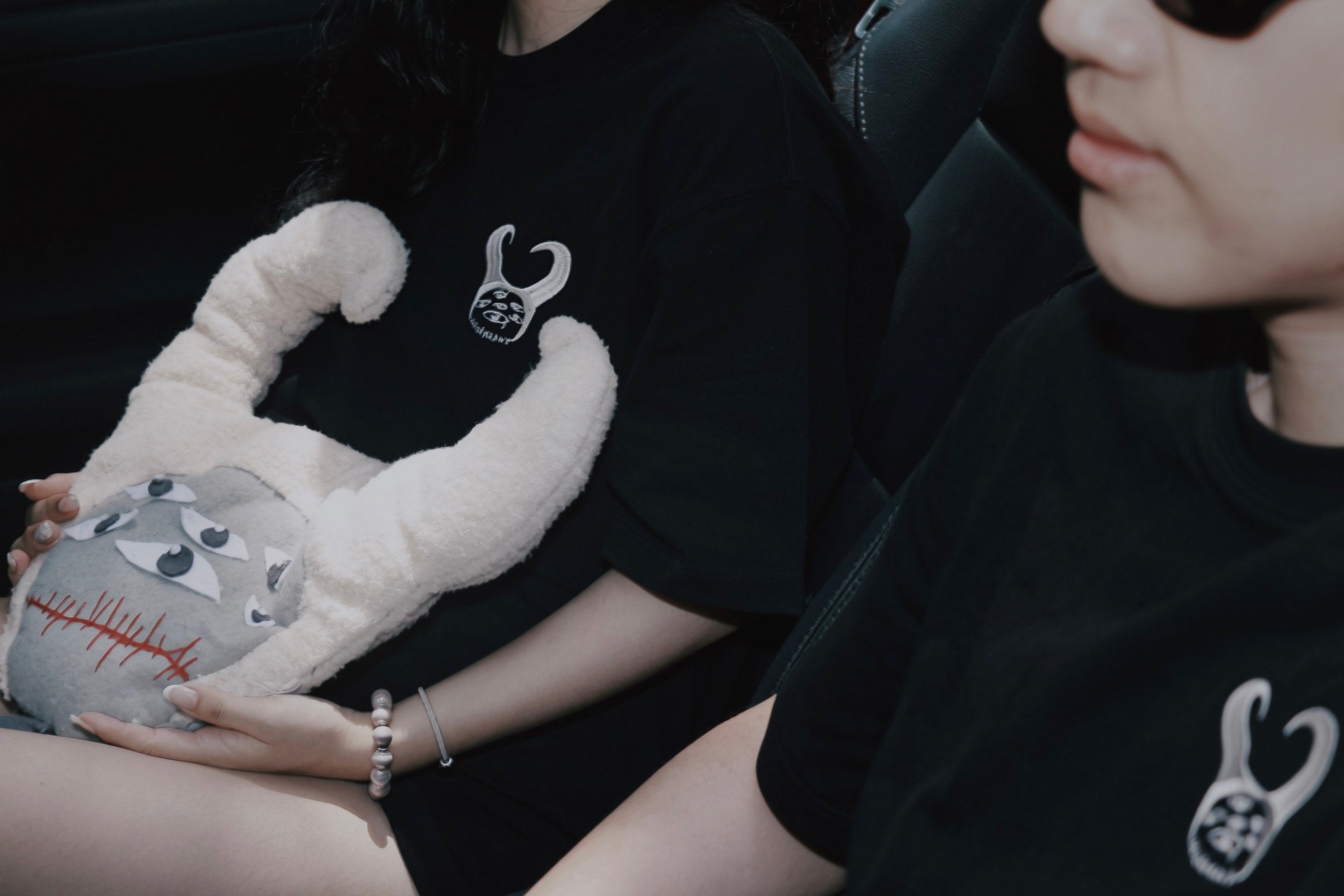 Two people wearing black LE GÉMEAUX T-shirts seated in a car, one holding a grey horned MON plush.