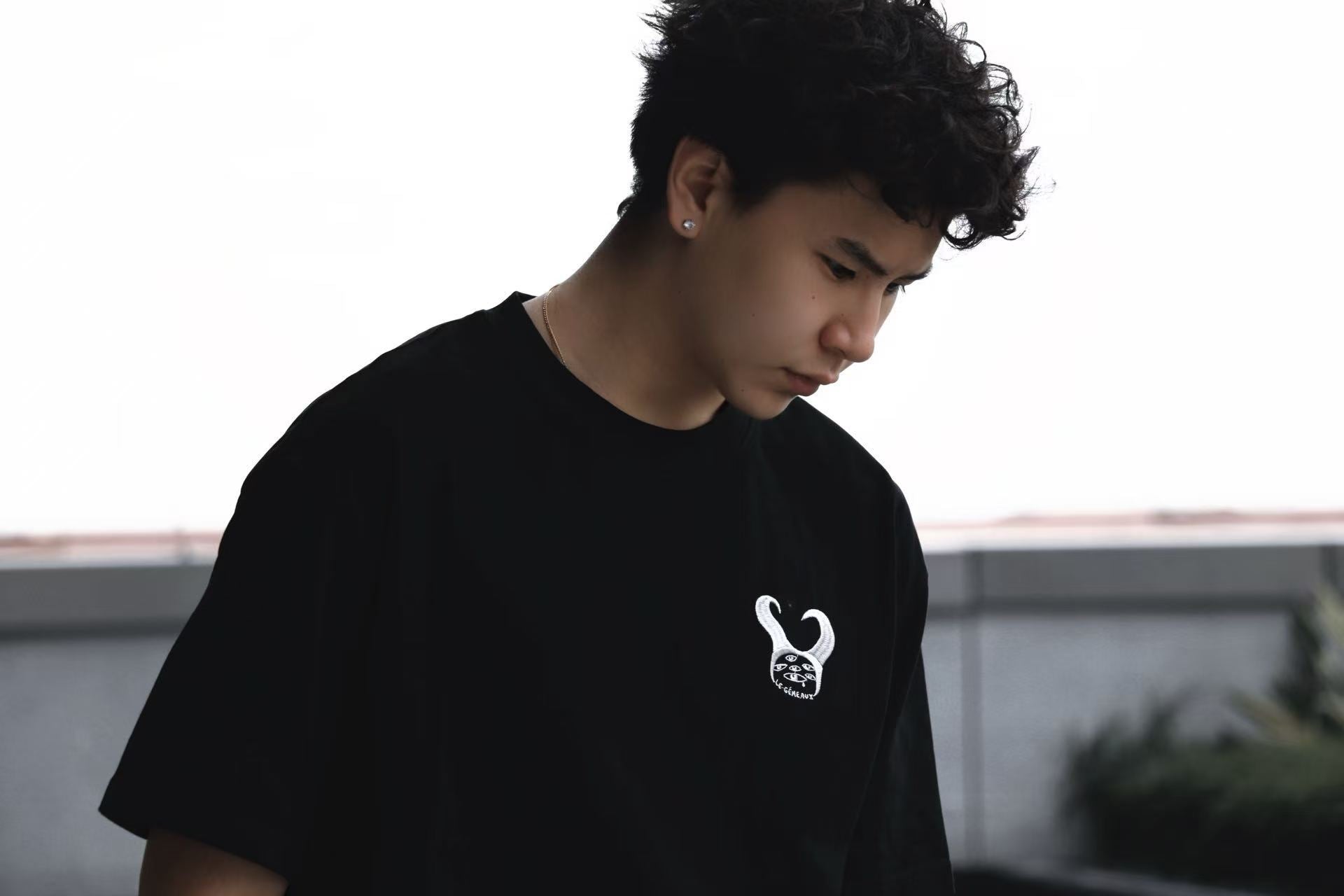 Person wearing a black LE GÉMEAUX T-shirt looking down outdoors.