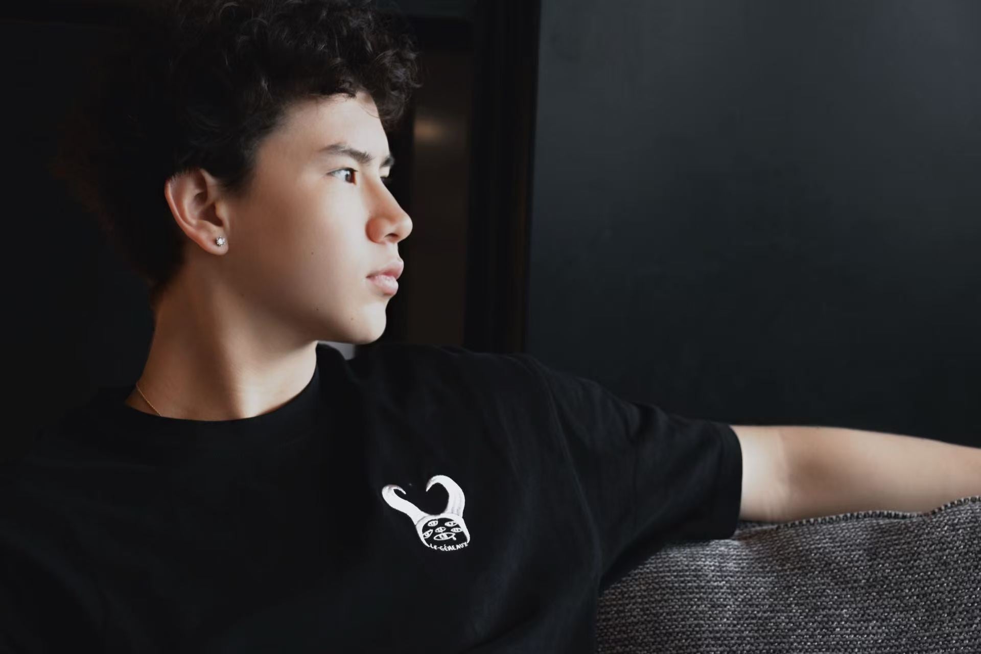 Person wearing a black graphic T-shirt, seated on a sofa and looking to the right.